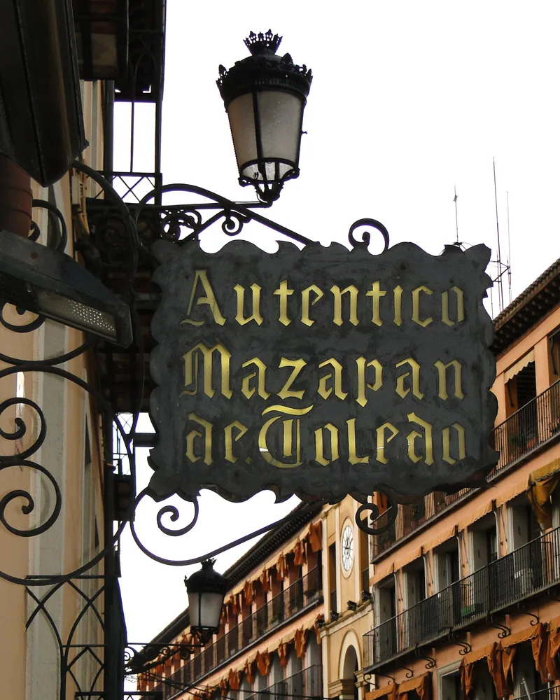 Cartel de forja con letras doradas que dice Auténtico Mazapán de Toledo, colgado sobre una calle de Toledo junto a un farol